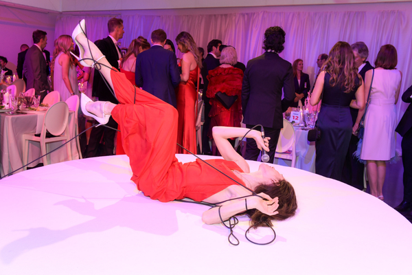 Performance artist James Knott lies on a elevated round white stage in a red dress and white heels, holding a microphone, with black cables wrapped around their arms and legs.