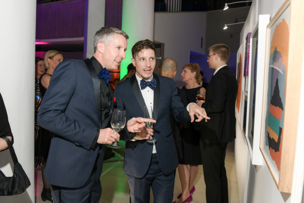 Two guests look and point at artwork that will be available in the live auction later in the evening, with other guests mingling in the background.
