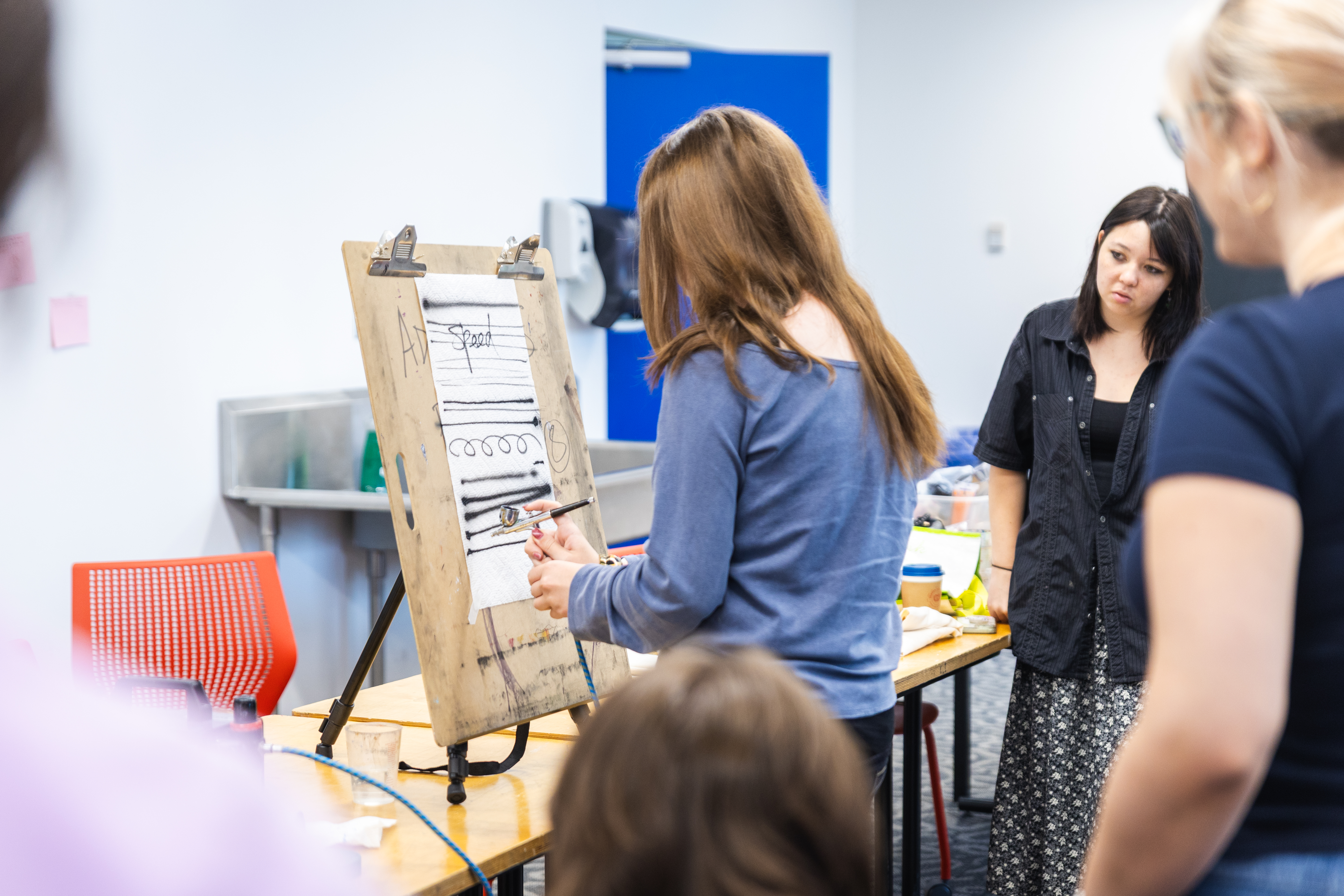 Student working at an easel on a table. Several other students are watching attentively. 