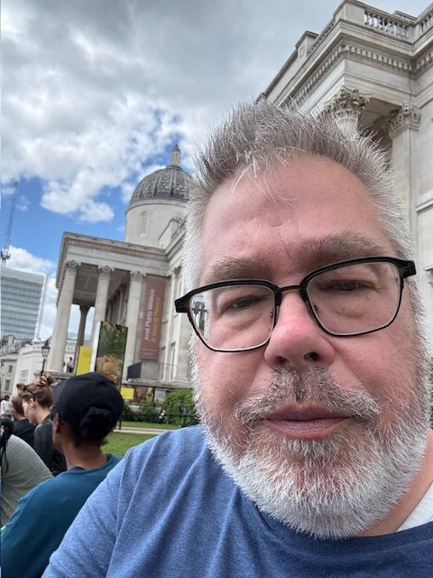 A man with grey hair and beard wearing black rimmed eye glasses and navy blue t-shirt.