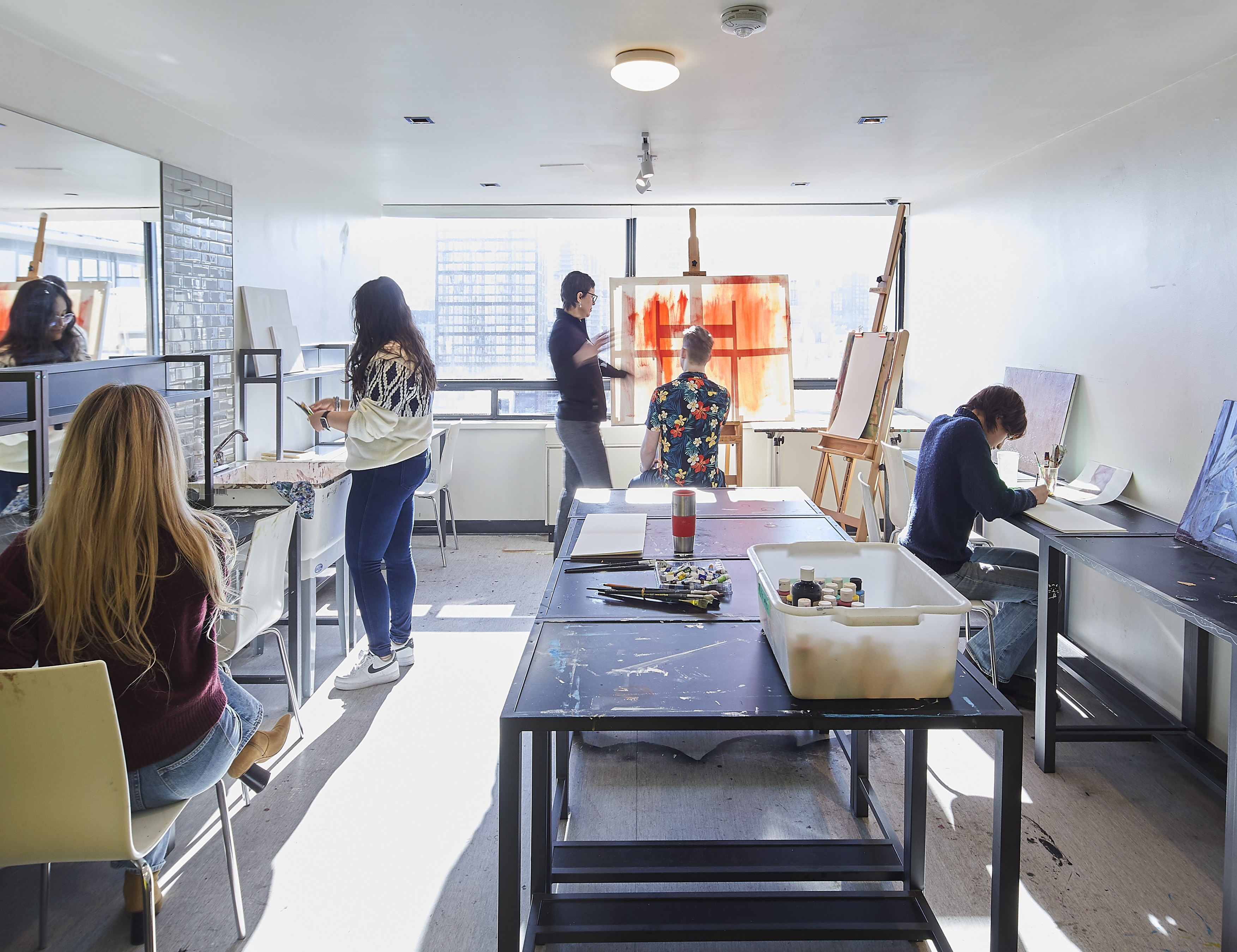 A group of students in a room with easals, paints and desks.