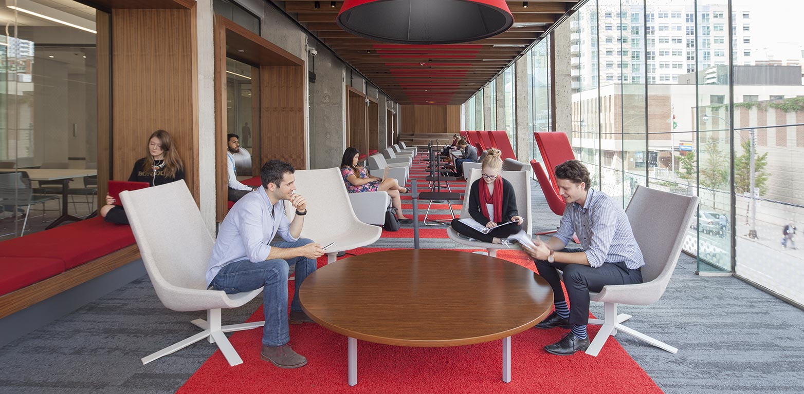 A room with windows along the right side with four students sitting in white chairs with red carpet, student off to the left side studying.