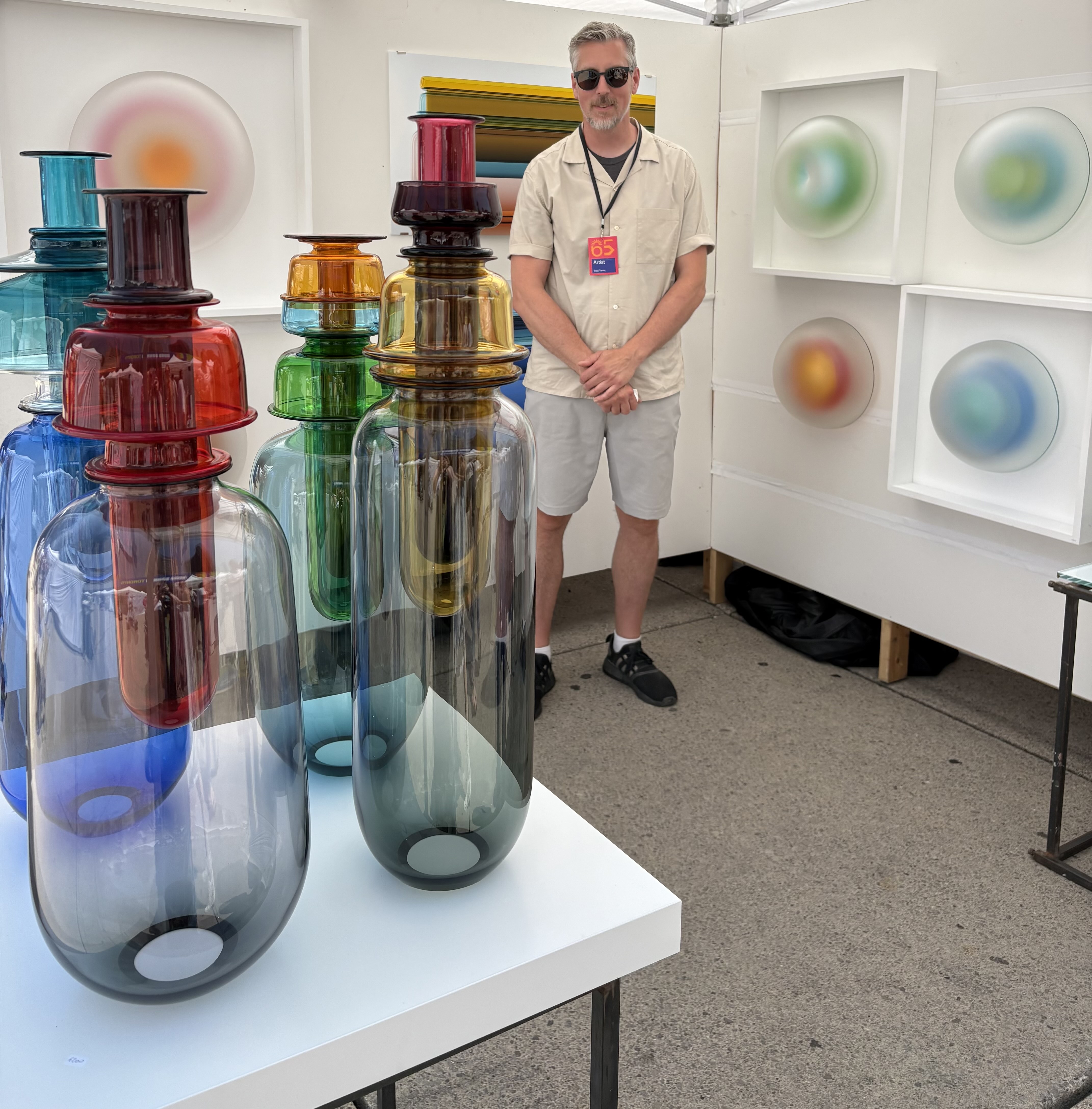 Man with short grey hair wearing sunglasses standing in his exhibit of glass work