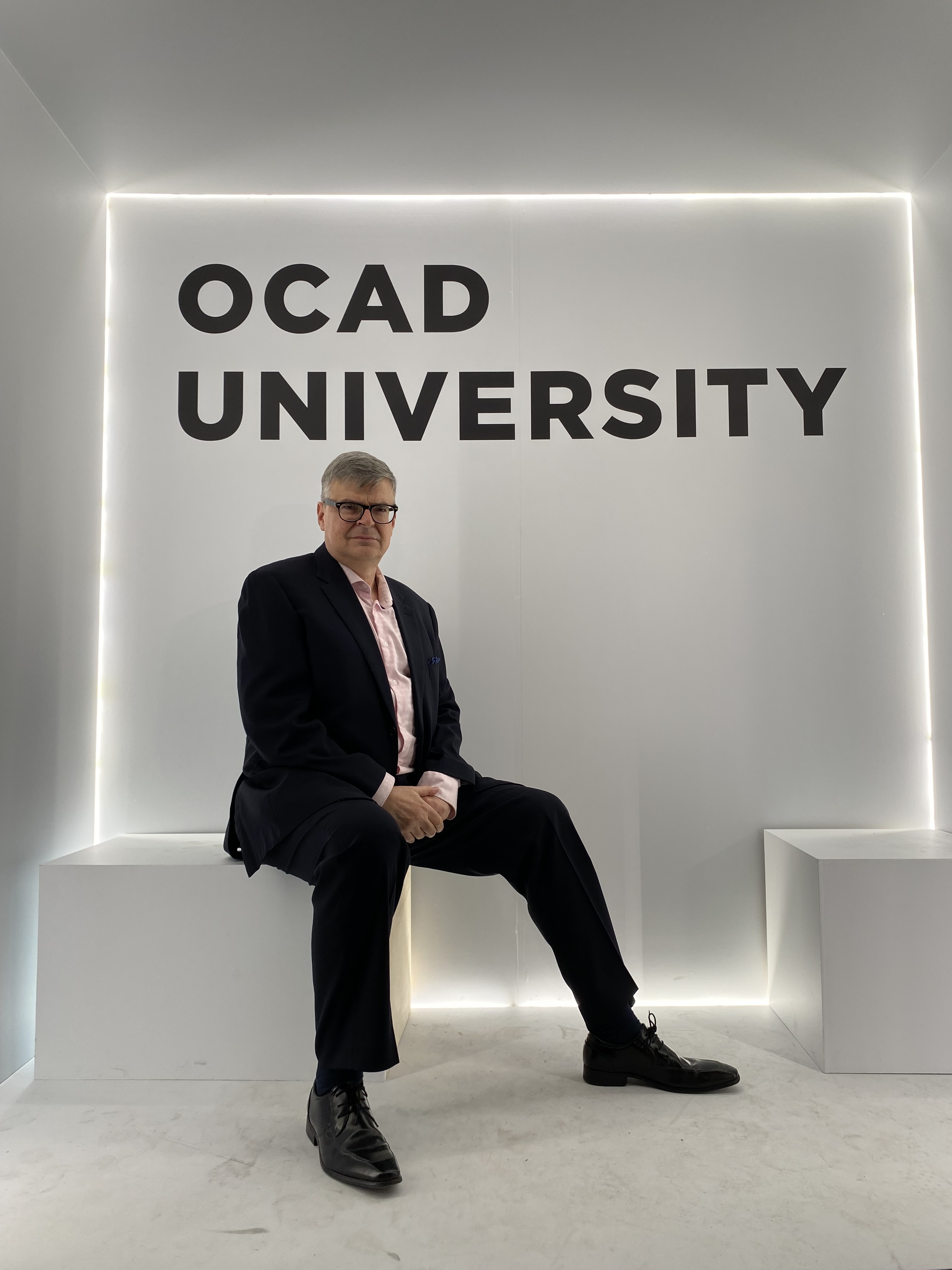 Man wearing classes wearing a black suite in front of a sign that says OCAD University