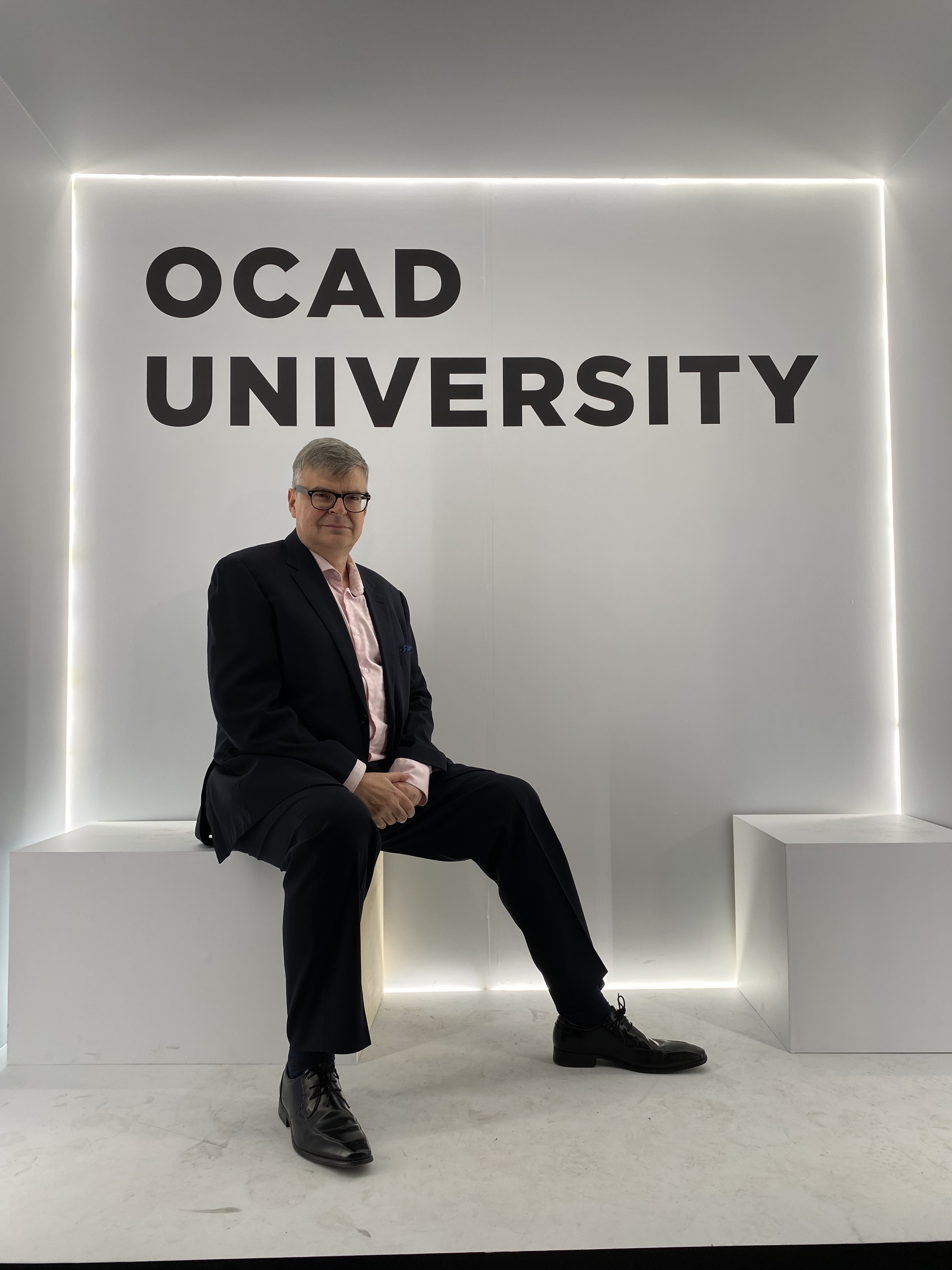 Man wearing black suit, white shirt and glasses sitting in box that says OCAD University