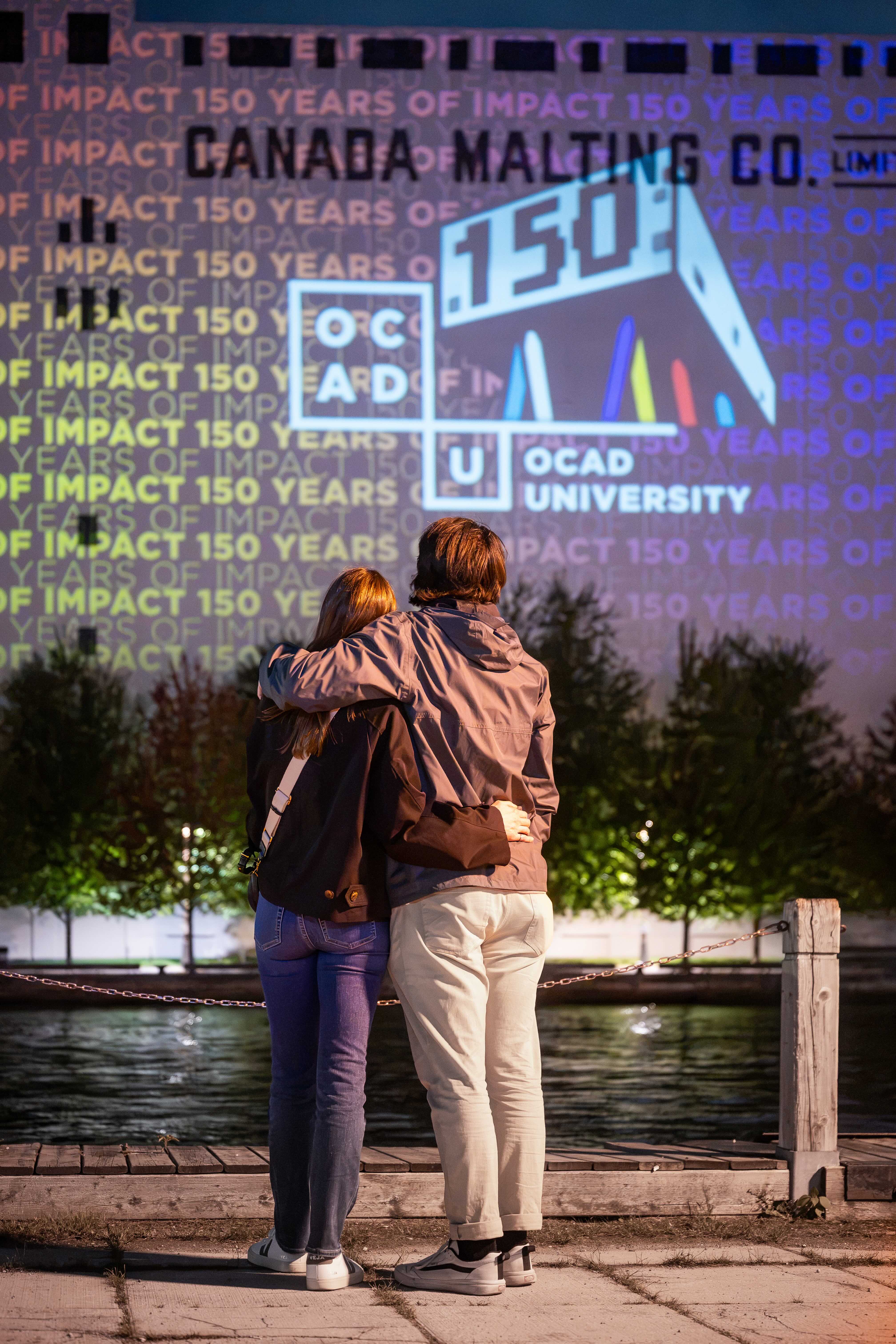two people watching a projection on Canada Malting Silos, 150 years of impact with OCAD U logo with 150th