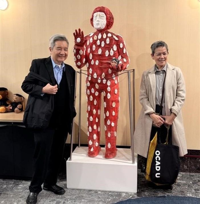 A man in a black suit and a woman in a beige coat with an art piece displayed between them. The art piece is a person that is wearing all red with white polka dots and red hair. 