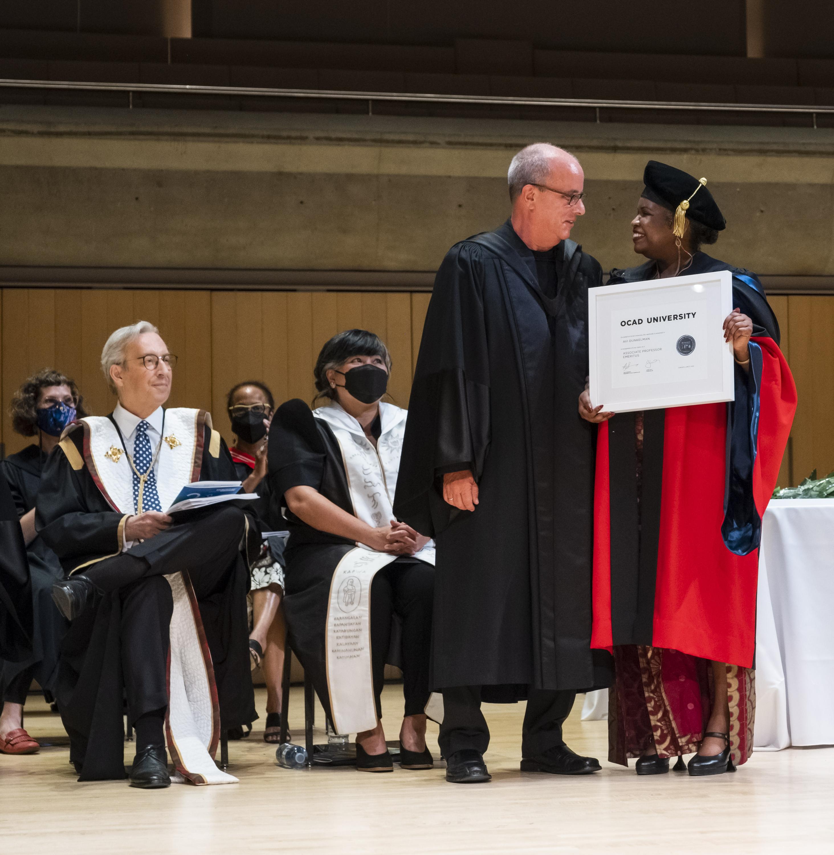 People in graduation regalia standing on a stage. A man is presented with a framed document.