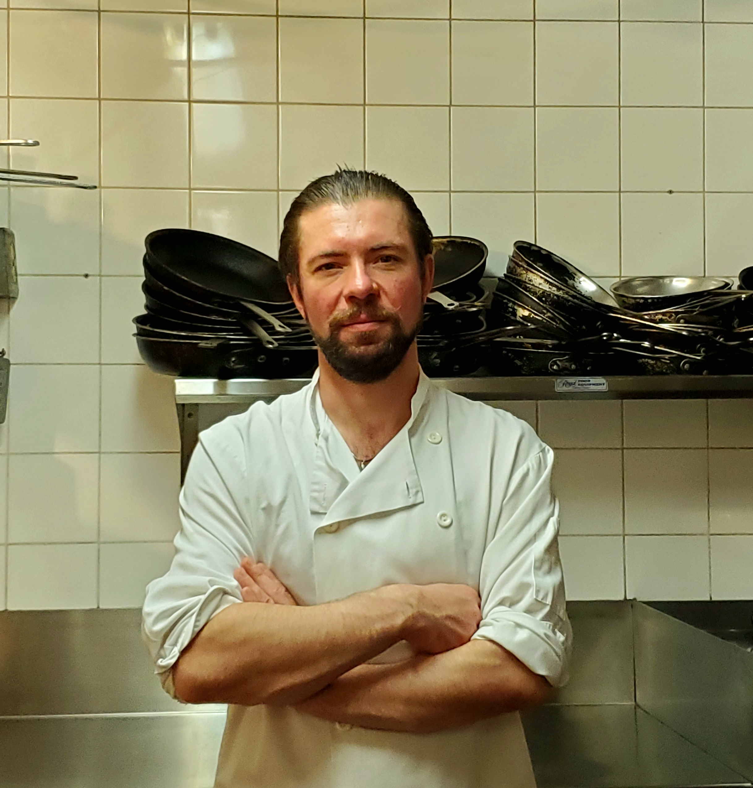A man with facial hair stands in a professional kitchen wearing a white chef's uniform.