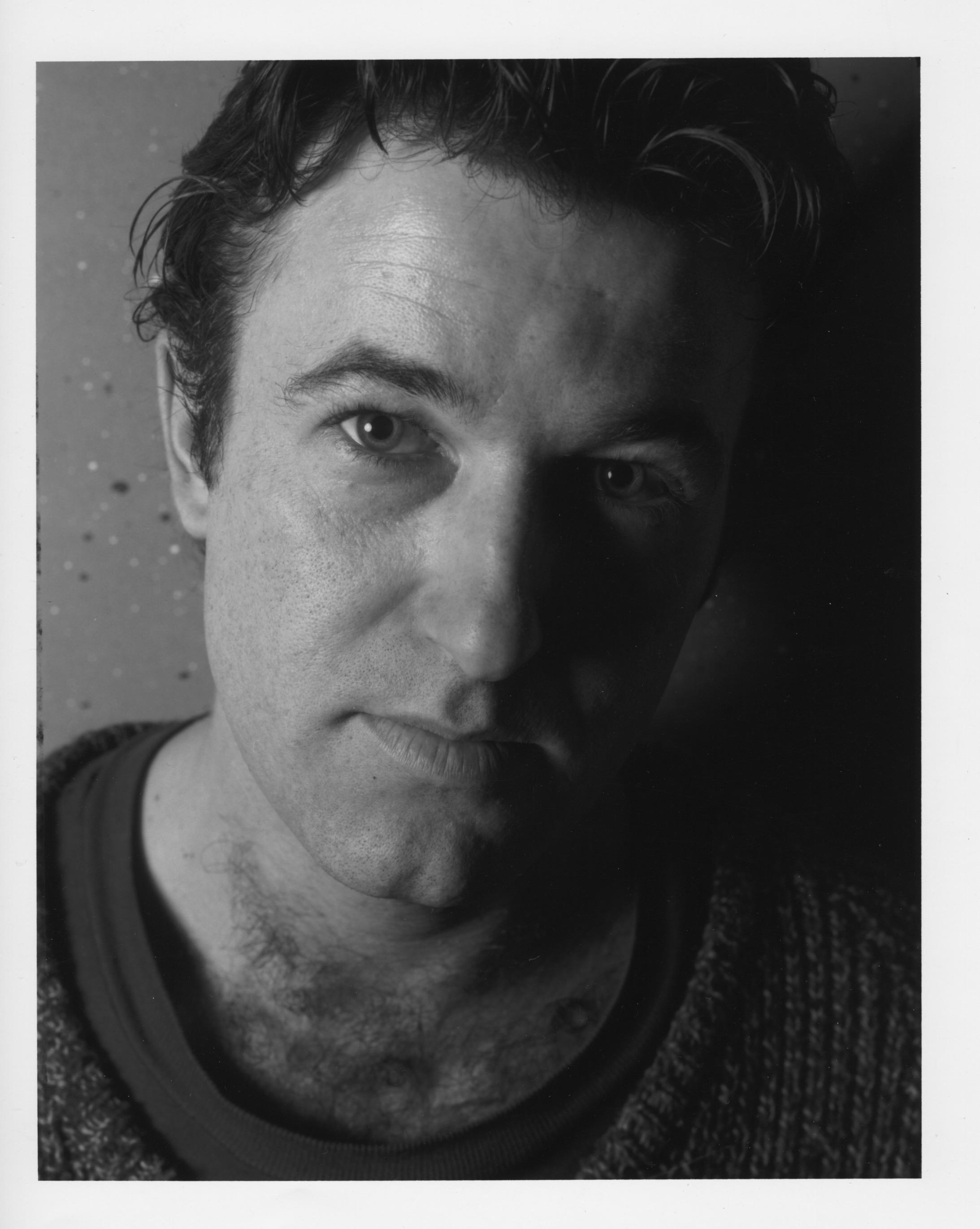 A black and white photo of a white man wearing a sweater looking directly into the camera.