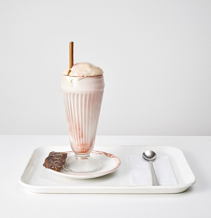 An ice cream float is visible in a glass on a white plastic tray with a metal spoon on a napkin to its right on a napkin.