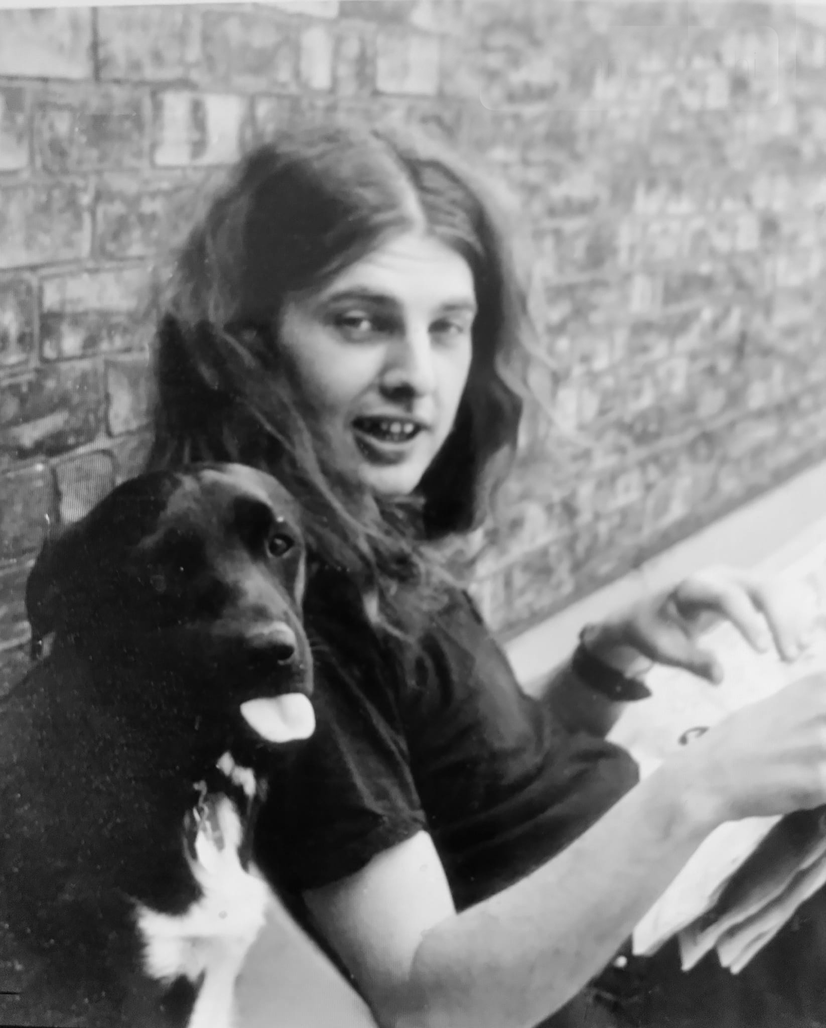 A black and white portrait of John Scott as a young person with long, wavy brown hair sitting beside a black dog with a brick wall behind.