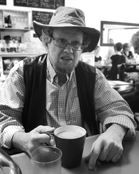 A photograph of a man with a wide brimmed hat and glasses sitting at a table in a cafe, holding the handle of a coffee mug.