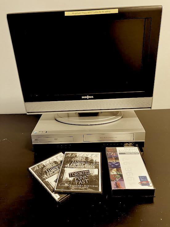 Image consists of a video monitor hooked up to a DVD/VHS player. Placed in front of the viewing station are two archival DVD’s and one archival VHS.