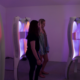 Two people observe a series of white sculptural works by Jane Tingley, Cindy Poremba and Marius Kintel in purple and pink hued lighting