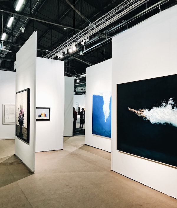 An image of an art fair, with booths and paintings on the walls. People are visible gathered in the background.