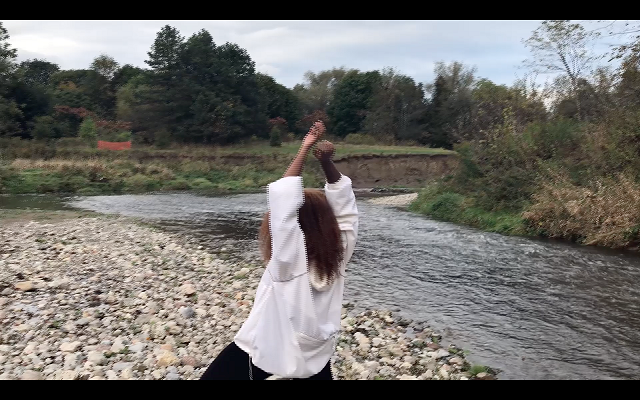 A person with long hair facing a river with their arms raised above their head, 