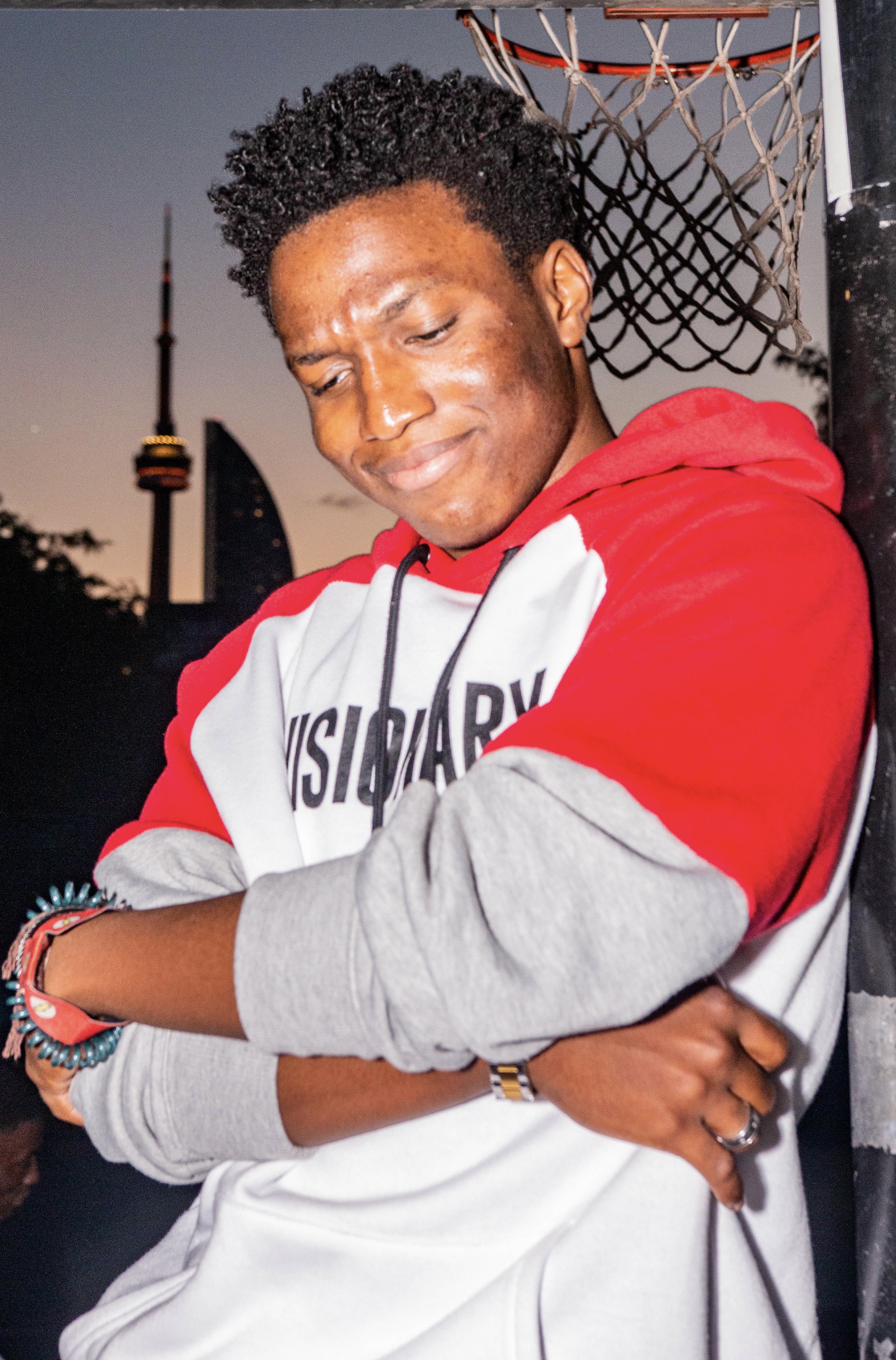 A photos of a young man standing under a basketball net with the CN tower in the background.