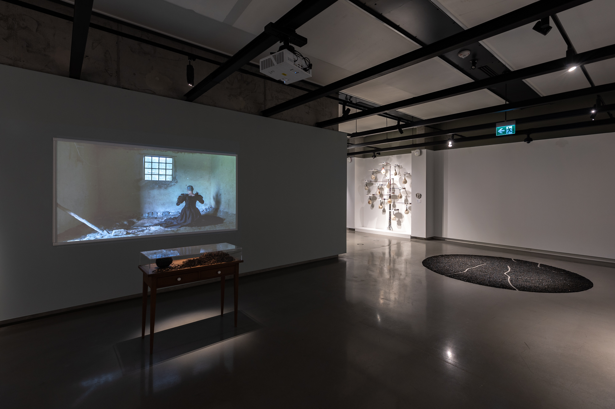 An overview of a exhibition. On the floor, there is an artwork of coal placed together as a large circle. There is a projection of a woman dressing black Victorian dress kneeling on the floor while weaving hair together. In front of the projection, there is display case of hair skein. On the wall, there is a multiple ceramic punching bags mounted. 