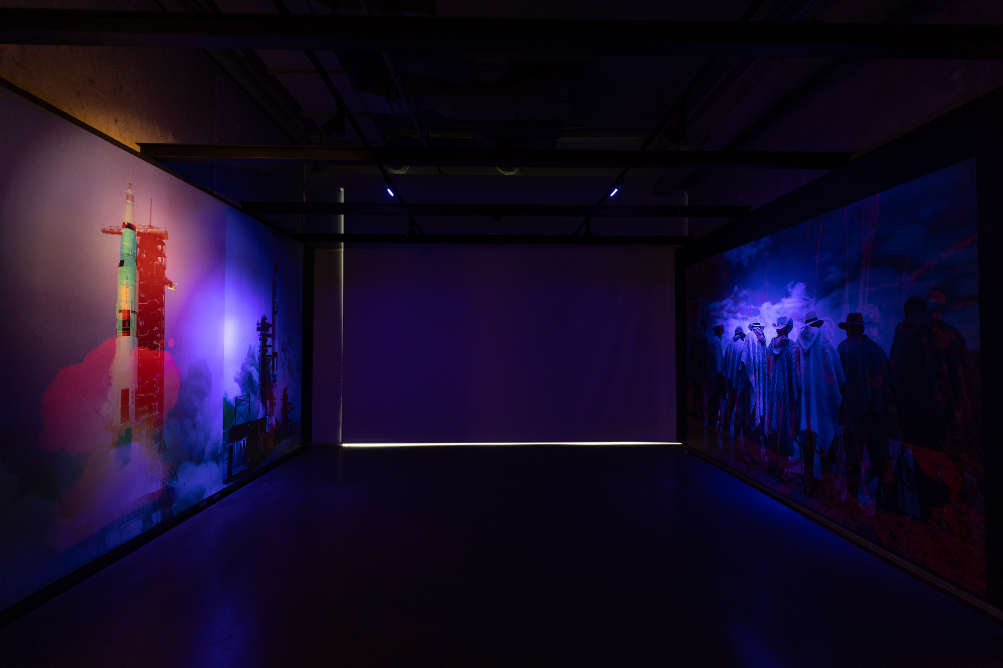 An image showing two archival images blown into large picture installed on two walls. On the left side is an image of a rocket launch. On the right side is an image of men wearing ponchos and hats.