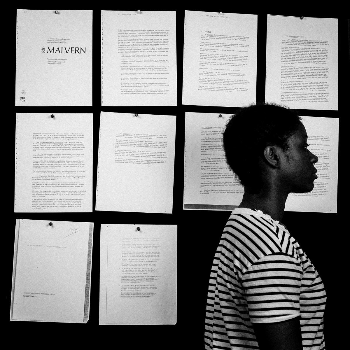 A black and white photo if a Black woman stands in front a grid of documents posted on a wall.
