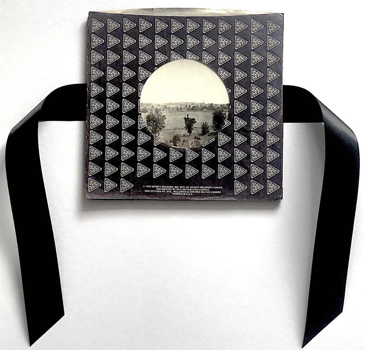 Artwork by Schem Rogerson Bader, A Storm is Coming From Paradise, 2024, assemblage, 10 x 10 inches. Black and white photograph in a record envelope, with black ribbons on either side, on a white background.