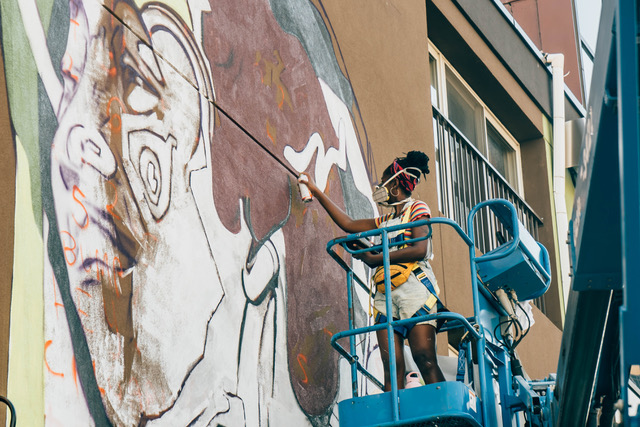 Curtia Wright wears a mask and stands on a lift, painting a large-scale mural of a face on the side of a building.