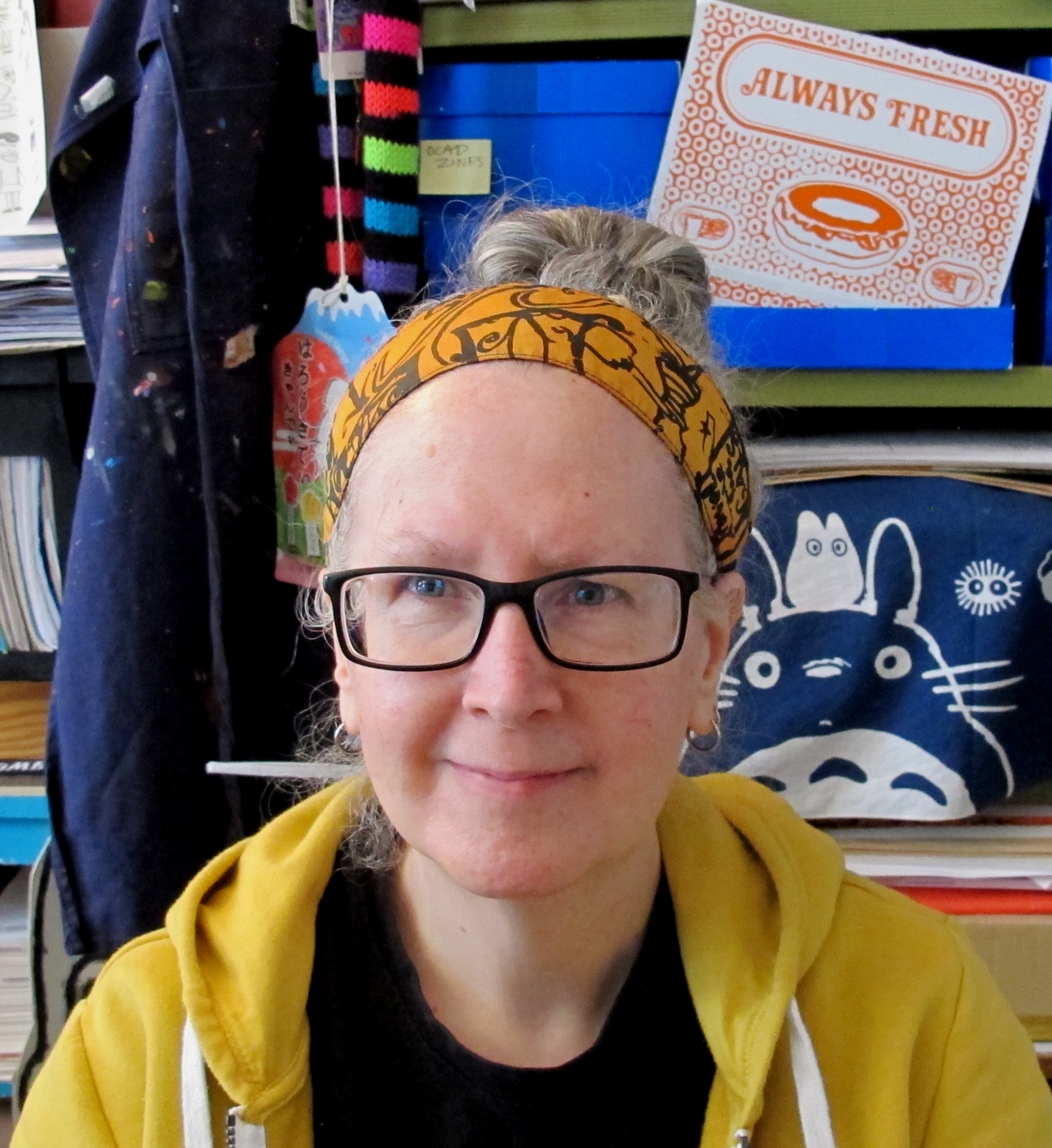 A woman with wearing a orange and black pattern head band, with grey hair pulled up into a bun. She is wearing black eyeglasses, has blue eyes and wearing a yellow hoodie with black t-shirt under