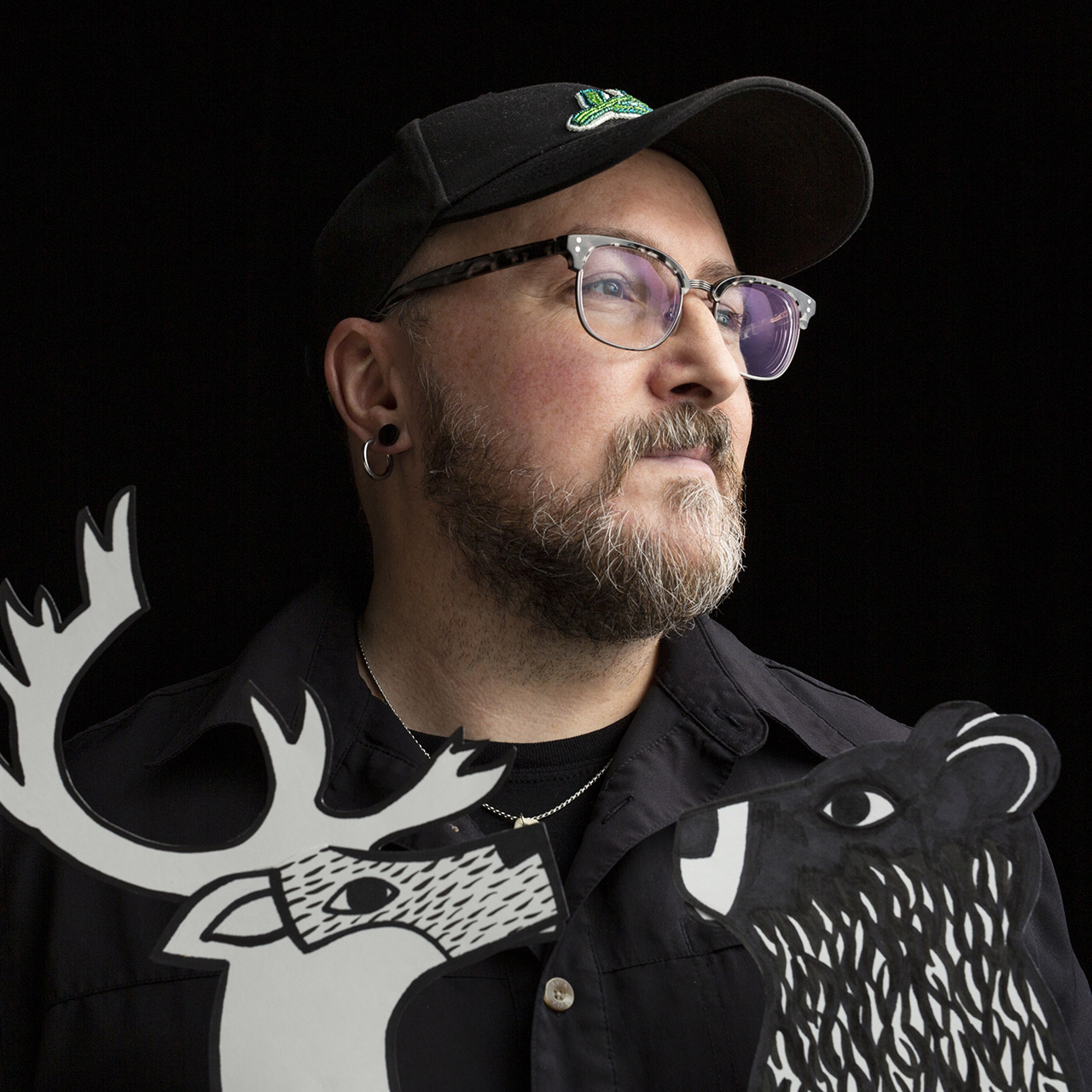 Portrait of Glenn Gear, wearing black hat, black shirt, and glasses, on a black background.