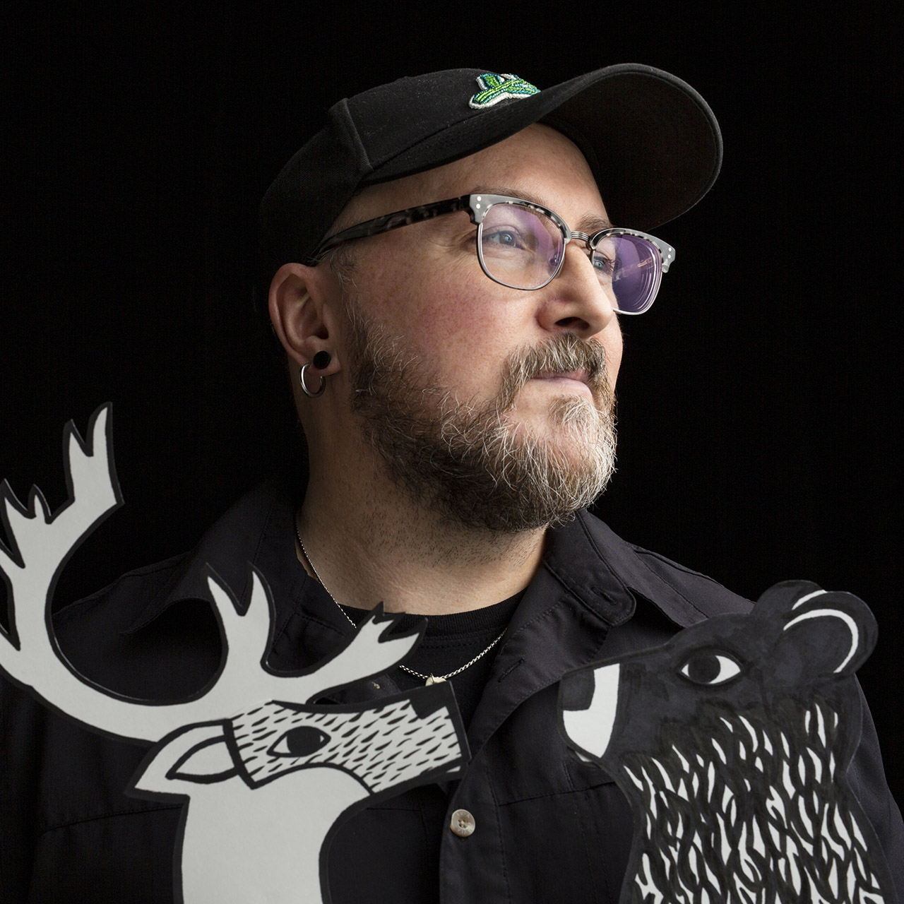 A portrait photograph of Glen Gear. He wears all black, a baseball cap, and rimmed glasses while facing 3/4 away from the camera. Two black and white illustrations of forest animals, a deer and a bear, are overlayed at the bottom of the photo