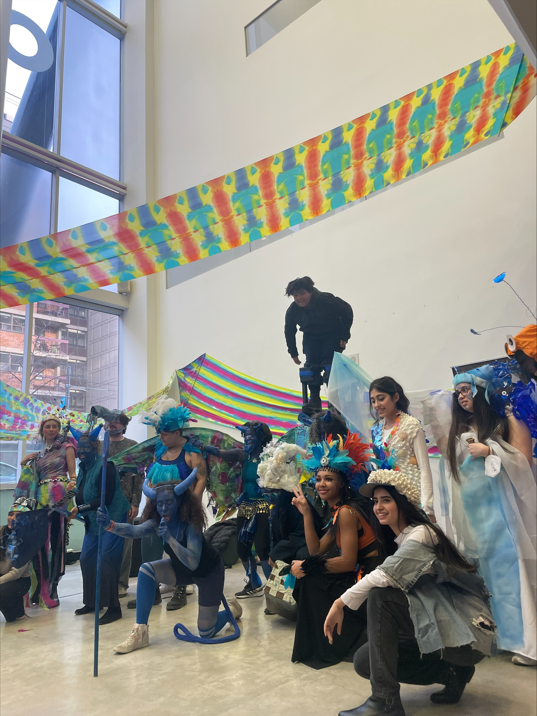 A group of participants in upcycled Mas costumes stand smiling in the lobby at 100 McCaul St. A colourful banner hangs above them. 