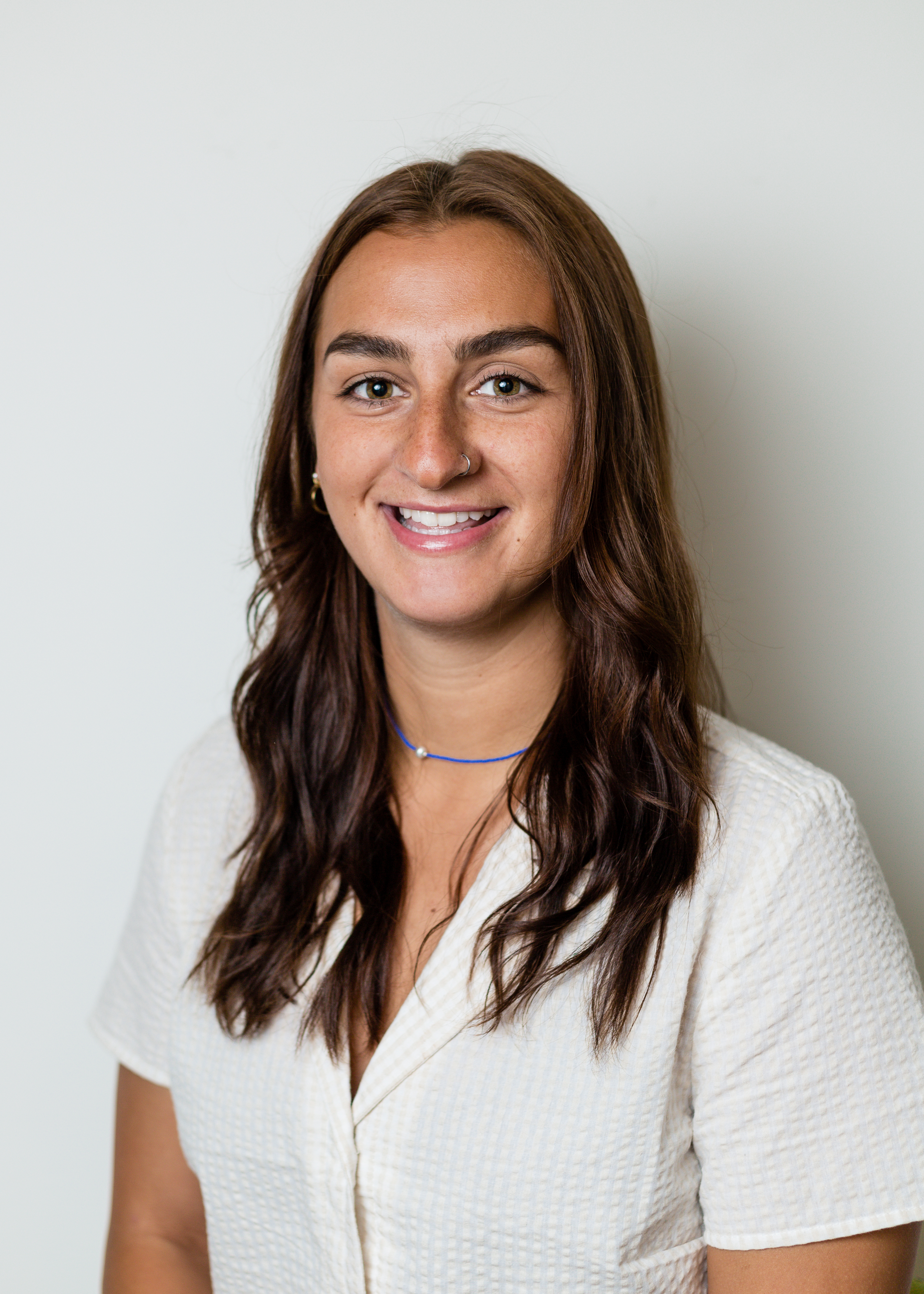 A woman with long brown hair with light brown eyes smiling at the camera wearing a short sleeve cream textured shirt.