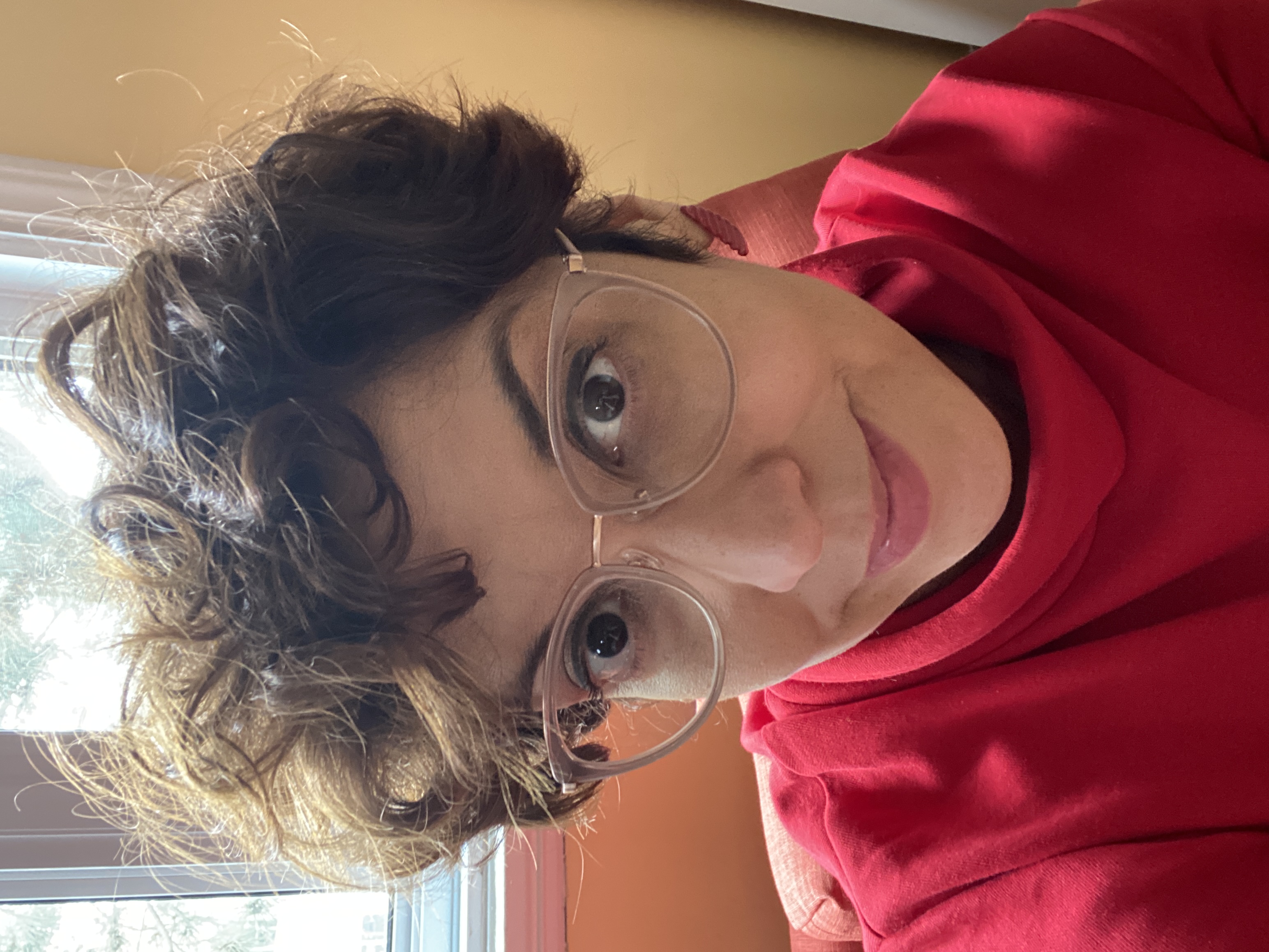 A woman wearing wire frame eye glass and red shirt with short brown curly hair smiling at the camera. 