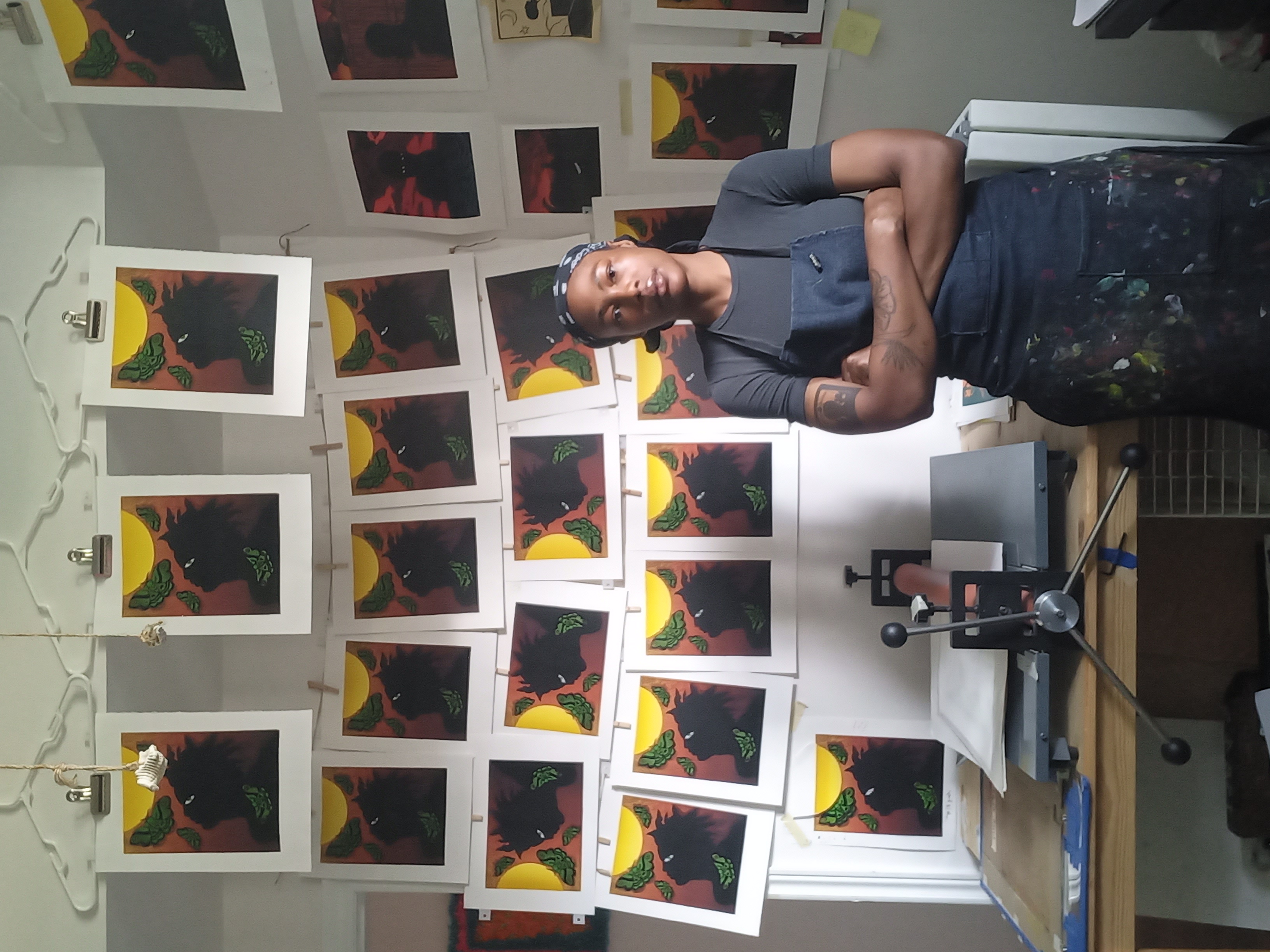 A full body image of a woman with arms crossed in front of her, wearing a black and white bandana, apron, black t-shirt. Behind her is her artwork, prints depicting a black person staring up in the sky, with a yellow sun and green leaves.