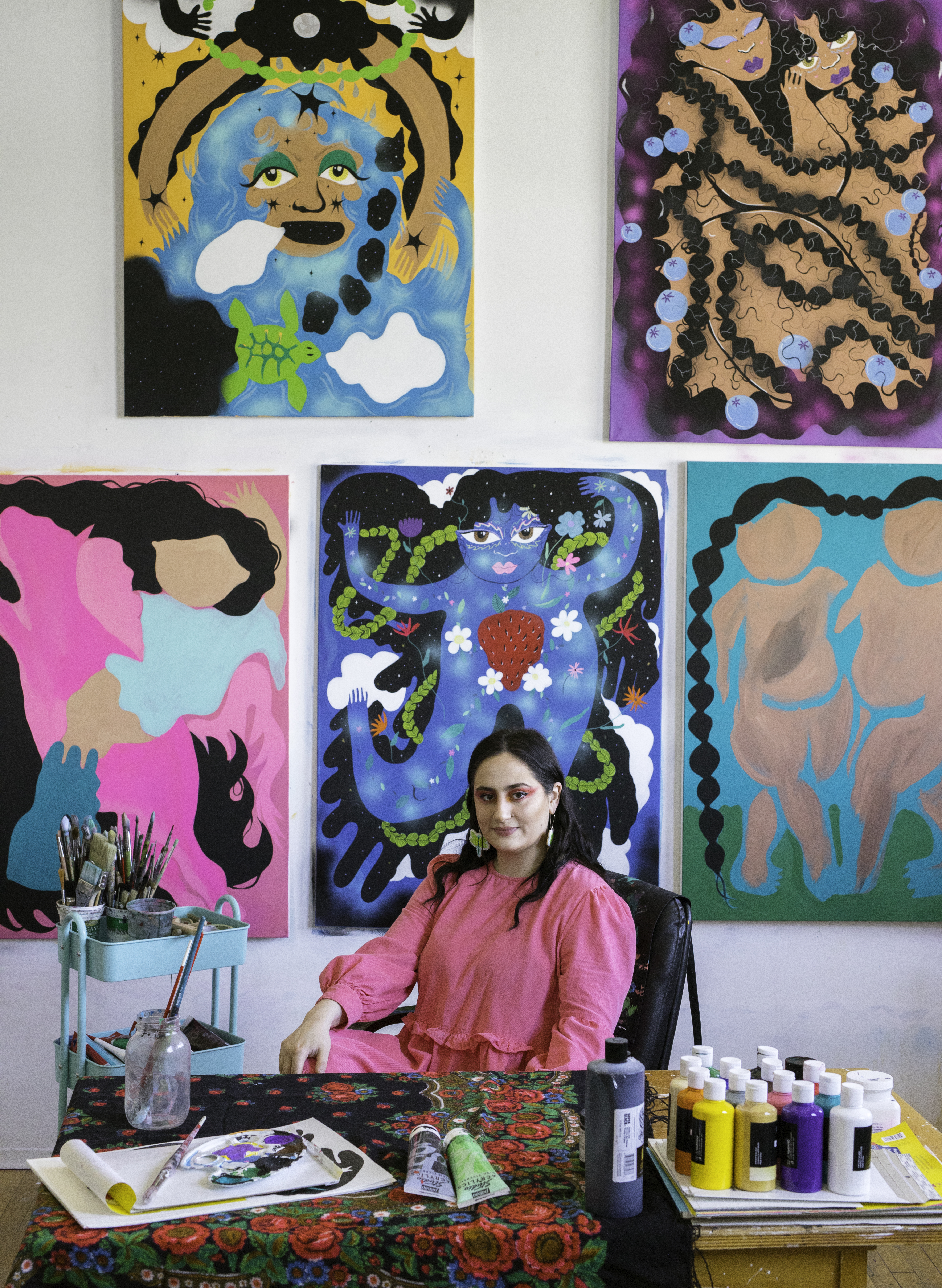 A person posing and surrounded by artwork. A woman with black hair, wearing hot pink dress.