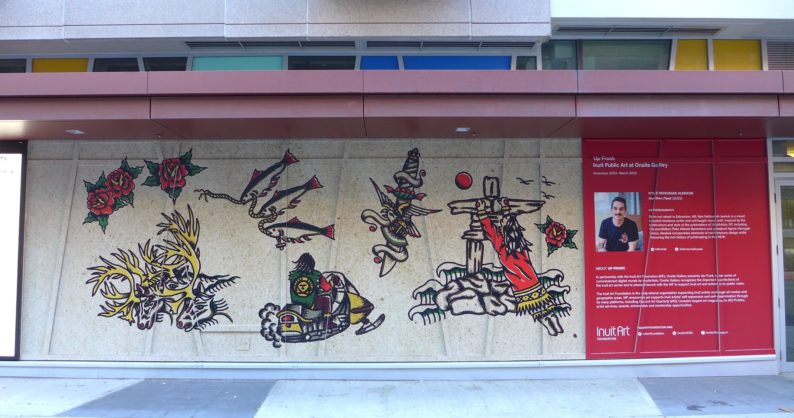Large mural that includes colourful artwork depicting a person on a skidoo, caribou herds, fishing lines, roses and inuksuk, all set against a sepia-toned background.