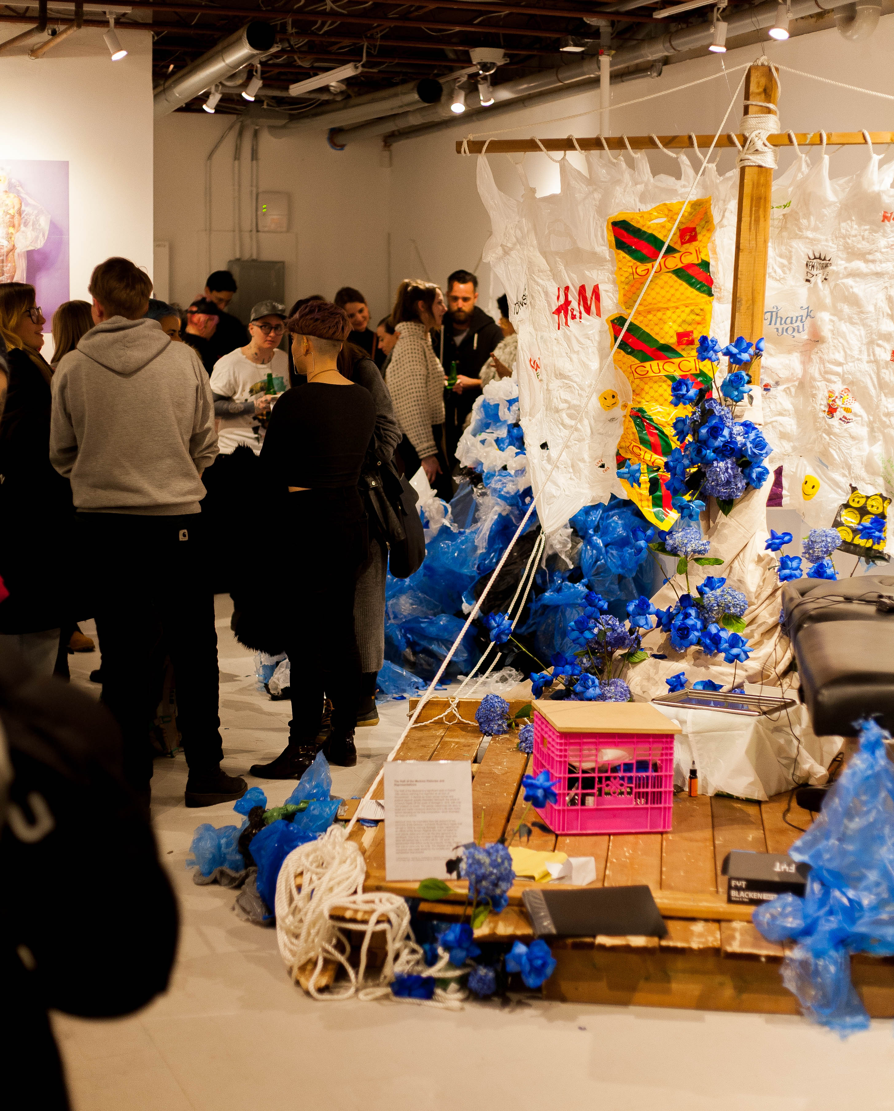 A photo of a busy gallery with a installation that resembles a raft with a sale, adorned with plastic bags.