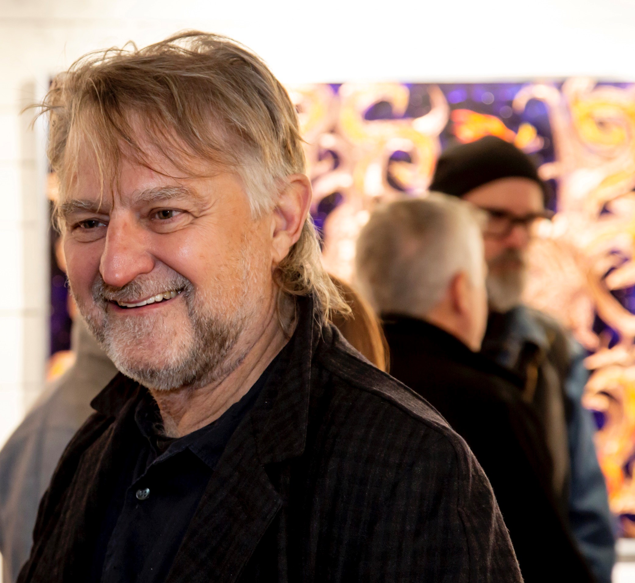 A headshot image of a older man with grey hair with a silver dark beard smiling at the camera. He is wearing a black shirt.