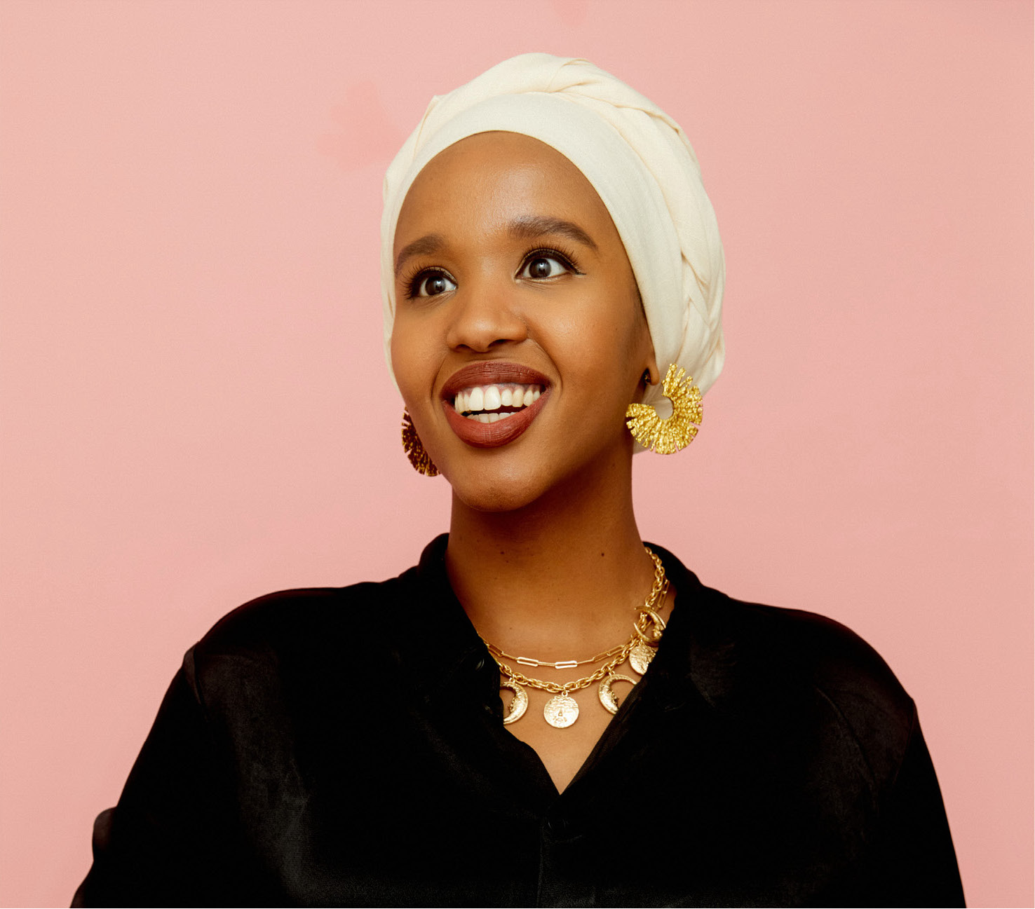 A woman wearing a cream color hijab with black satin long sleeve shirt posing to smile for the camera