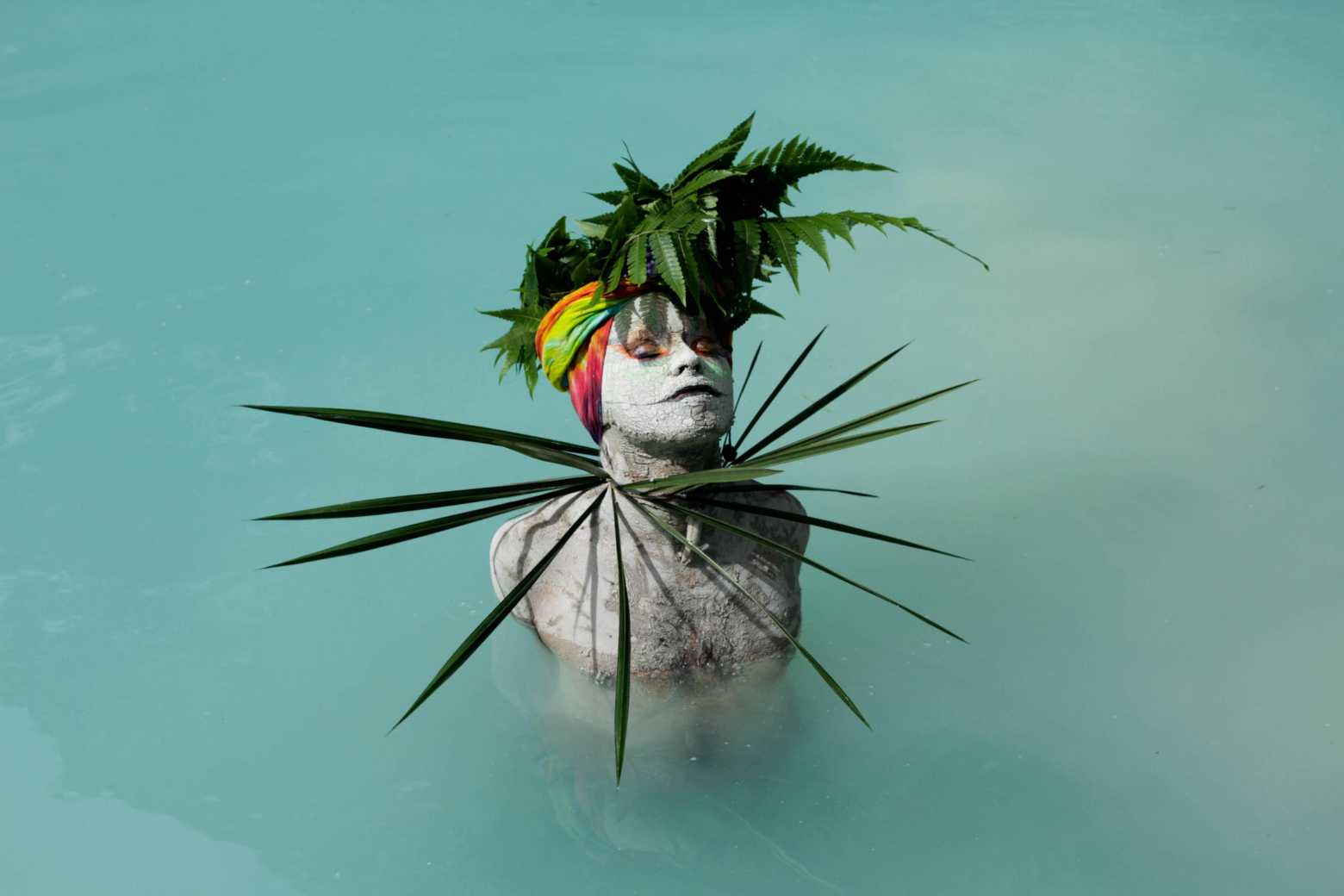 A photo of a person half submerged in a body of water with their eyes closed and skin painted white wearing head and neck pieces made of green foliage and a tie-dye head covering.