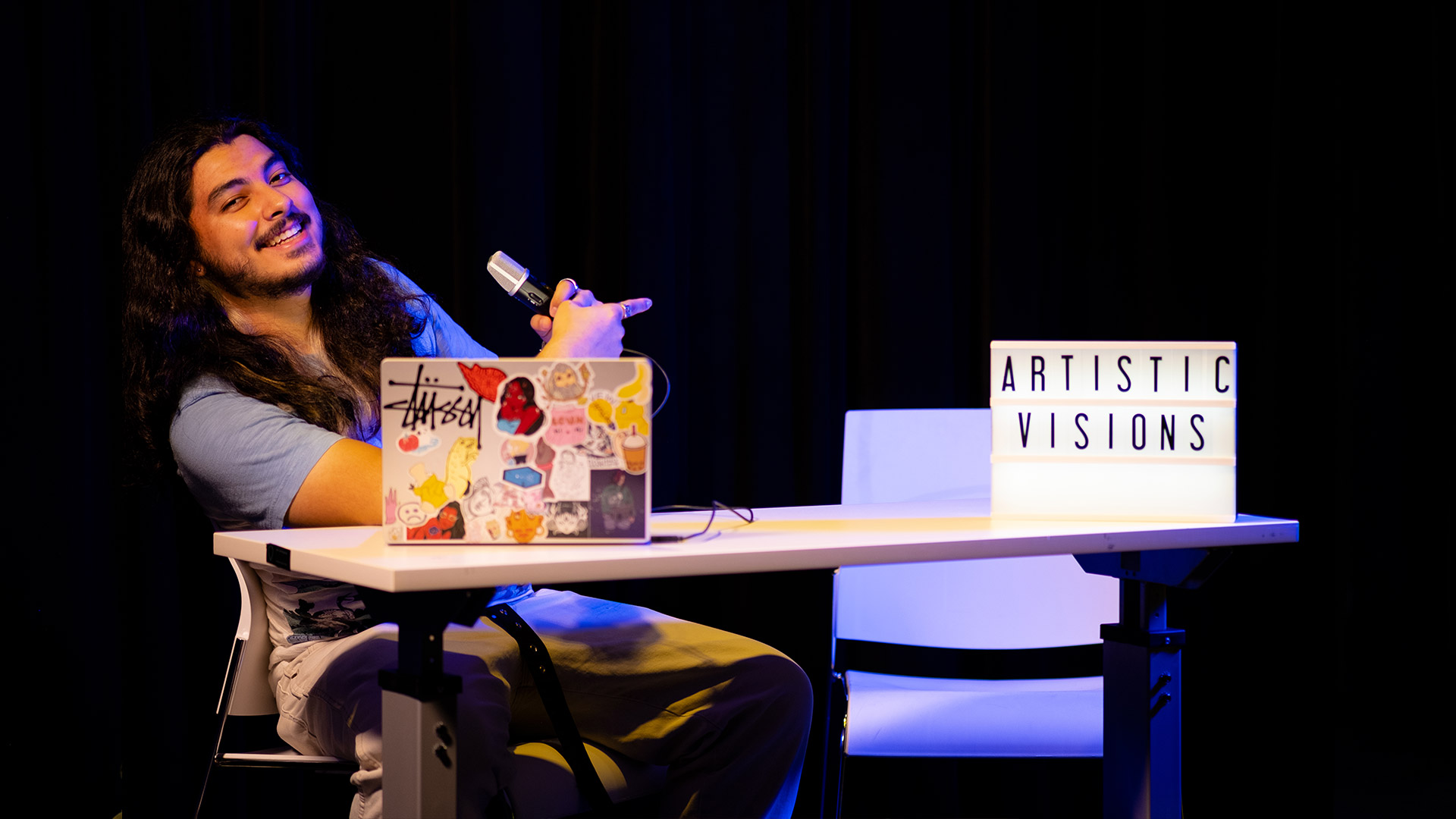 A photo of a young man with a beard sitting at a desk with a laptop and a sing that says Artistic Visions.