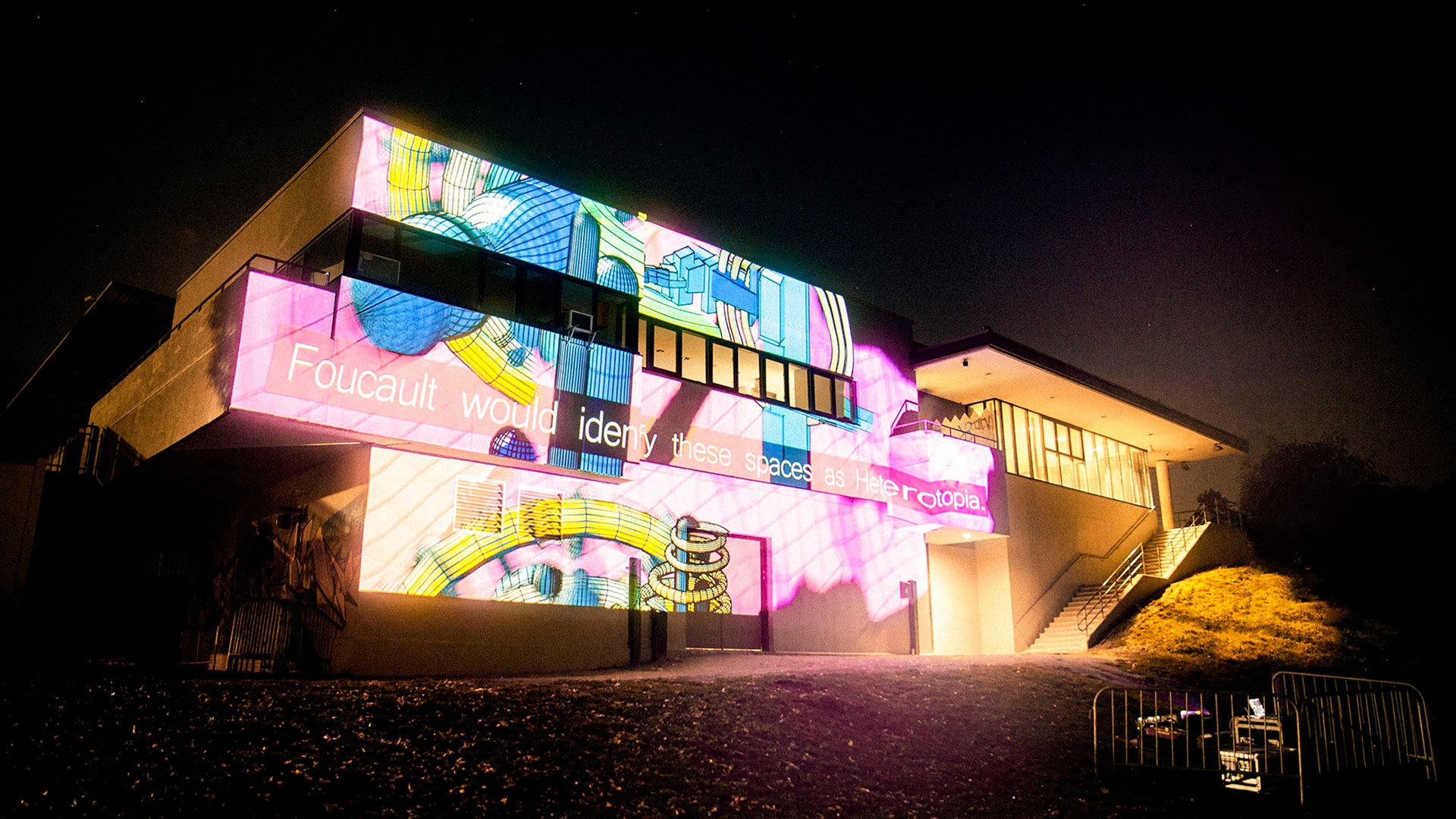 A photo of a building. On the wall are digital projections of colourful art and the text that reads "Foulcart would identify these spaces as heterotopia"