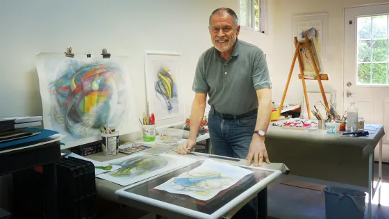 A photo of a smiling man wearing a greent shirt and blue jeans. He is standing in front of artprints and beside an easel in an art studio.