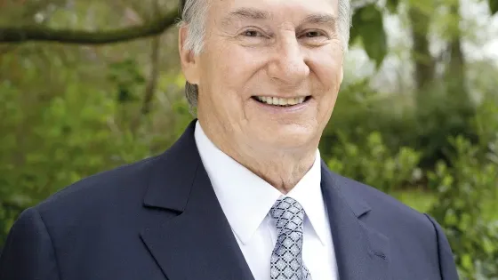 Photo of The Aga Khan wearing a navy suit , white shirt and blue and white patterned tie. 
