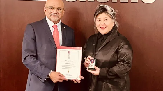 At left, man in a grey suit and red tie holding a certificate with the the city of Toronto in the background and the words, Toronto, in the background; at right, a woman with grey and black hair wearing a black jacket and pants holding a medal