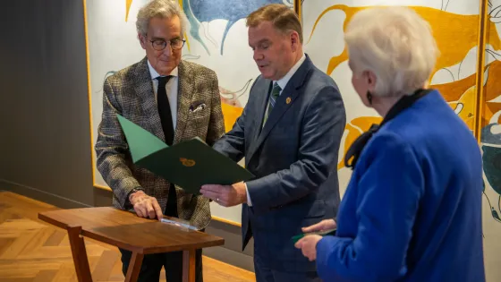 Three people in photo, at left a man with white hair and glasses wearing a grey patterned suit, man in middle holding a certificate and at far right woman with white hair wearing blue suit