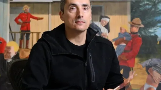 Colour photo of Kent Monkman holding a paintbrush, seated in front of his art work. 