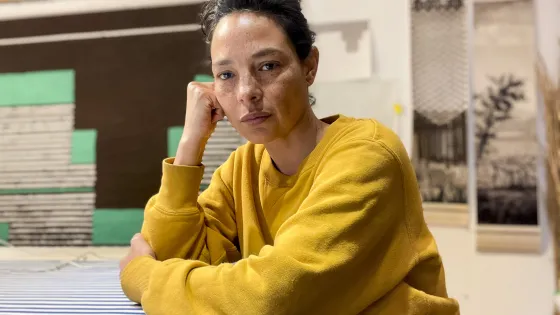 Photo of Christine Howard Sandoval sitting at a table, wearing a yellow sweater and with her arms on the tabletop.