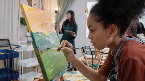 Young female teen is painting at an easel and an instructor is standing in the background.