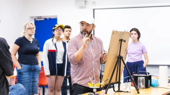 Students gather around an instructor giving an art demonstration in a studio space. Attention is focussed around an easel placed on top of a table. 